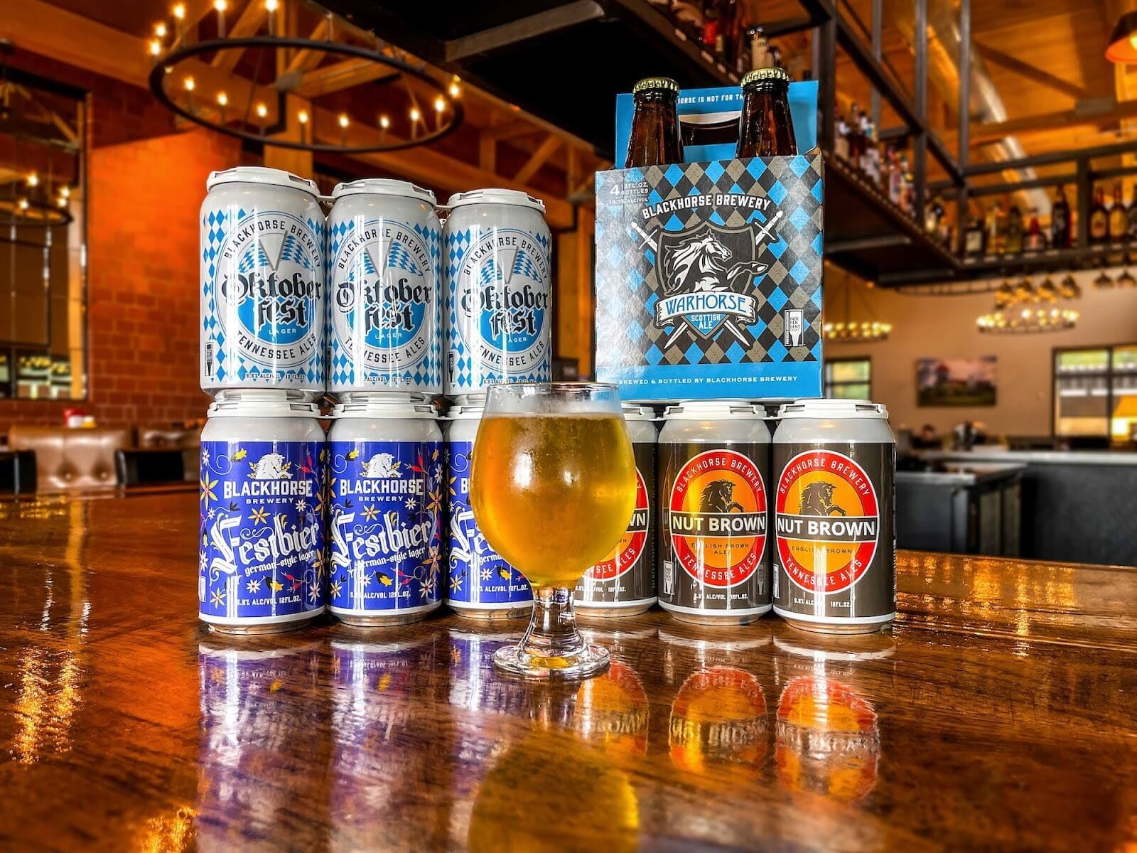 Cans of beer stacked together and a glass of beer in front of them on top of a table.